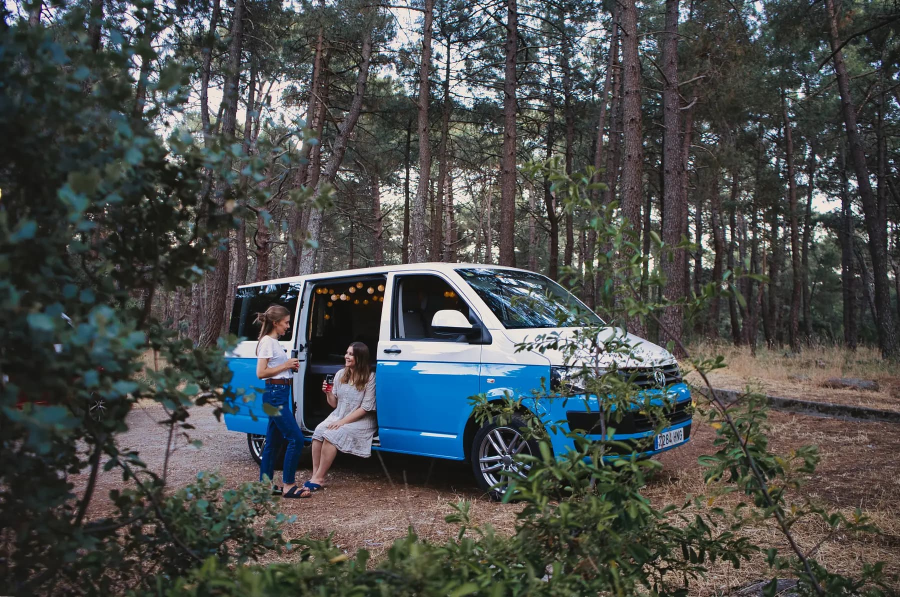 Camper beWinkle azul aparcada en un pinar con dos viajeras charlando